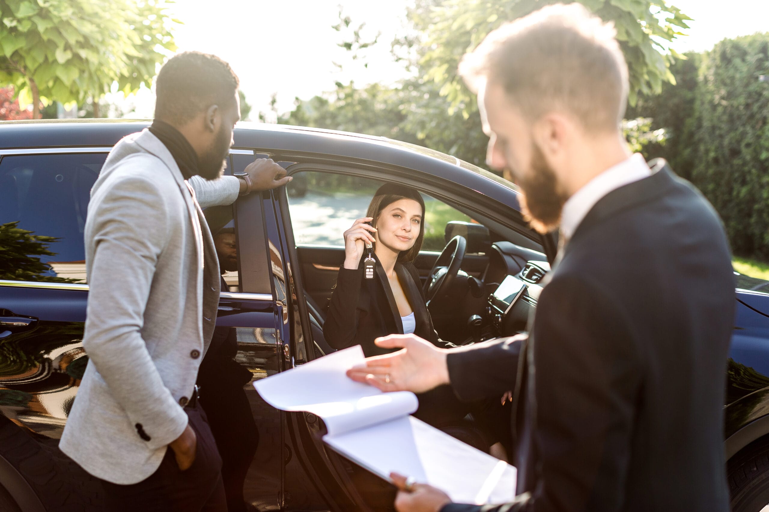 Happy multiethnical couple, African man and Caucasian woman, buying the car, black crossover, woman is sitting in the car and holding car keys. Young salesman holds folder with contract for sale
