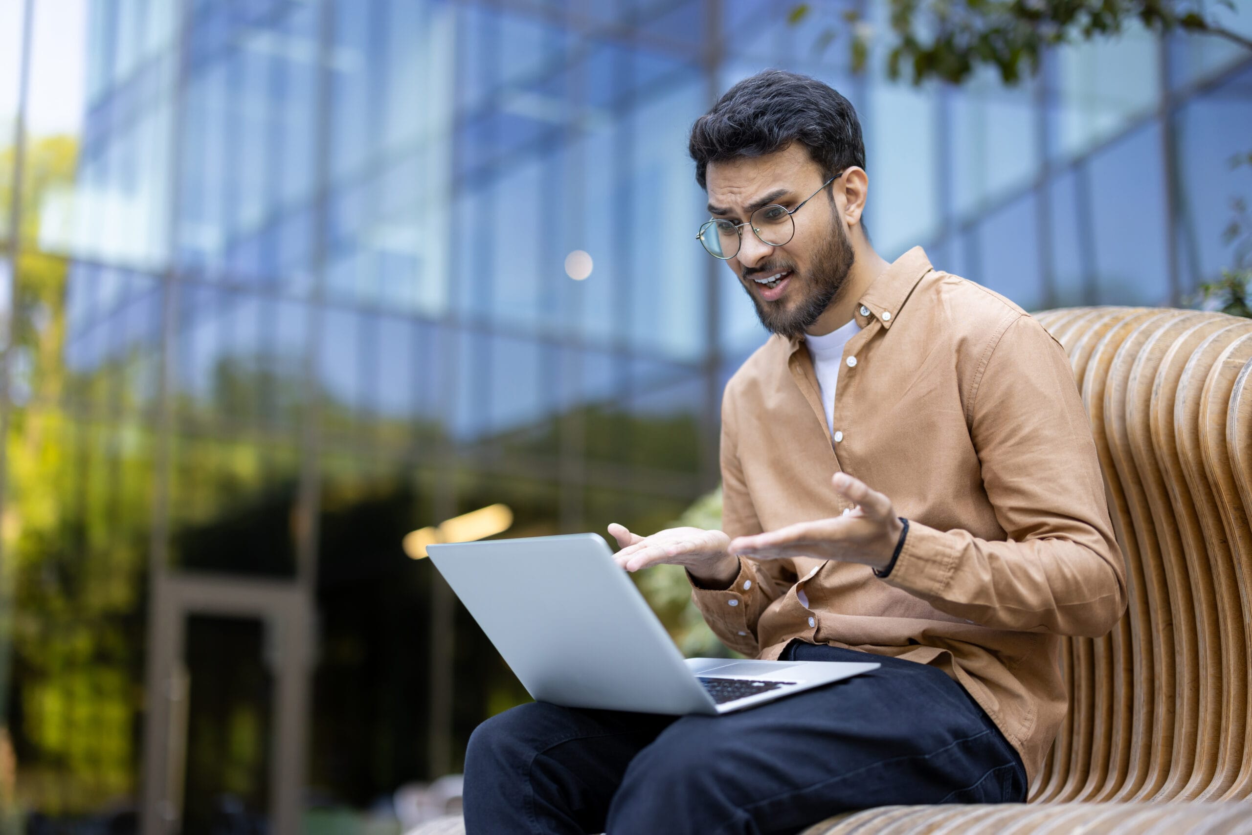 Young man sitting outdoors, looking frustrated while using laptop, expressing confusion, stress, and dissatisfaction during remote work on a sunny day